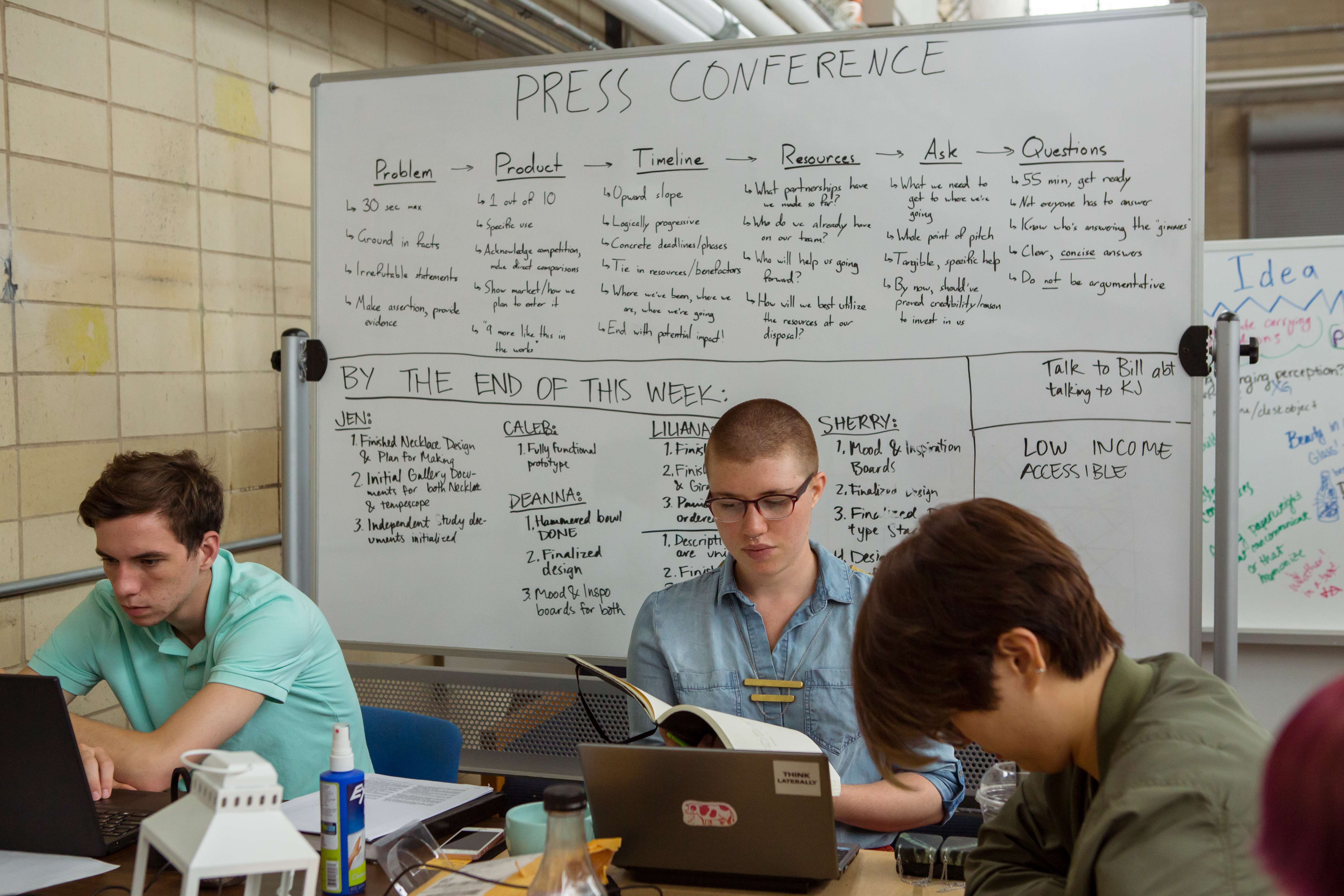 Three people are seated at a table, working. Behind them is a whiteboard with written goals for the week and points to keep in mind.