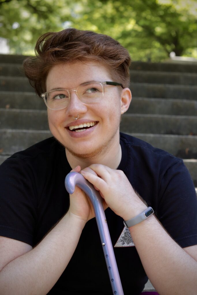 Photo portrait of a young individual with short auburn hair wearing a black T-shirt and clear acrylic glasses. They are smiling and holding a purple cane.